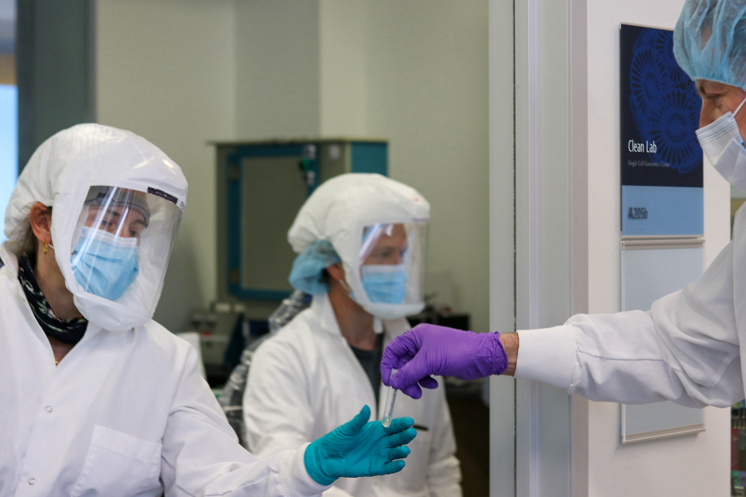 Three scientists in lab wearing protective equipment.  Scientist hands test tube to another scientist.